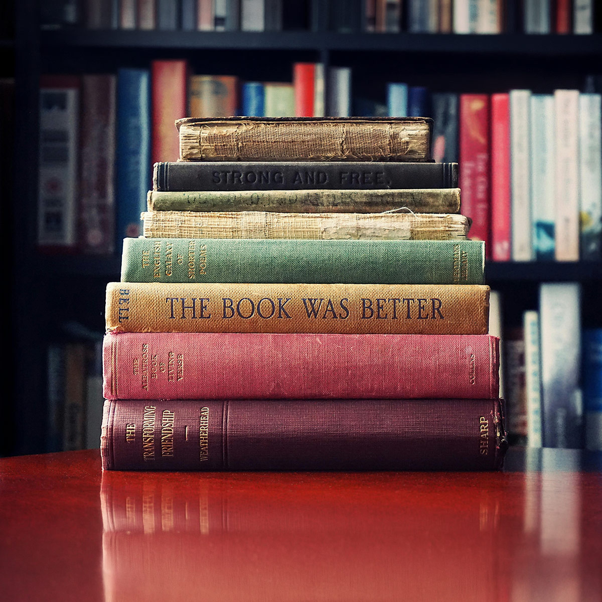 Stapel alter Bücher auf einem Tisch vor einem Bücherregal; auf dem Buchrücken eines Bandes steht „The Book Was Better“.
