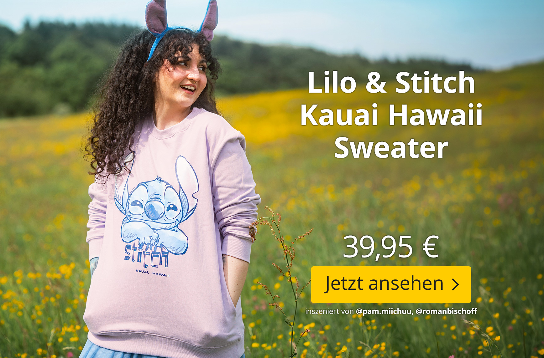 Person mit lockigen Haaren und Stitch-Ohren-Haarreif steht auf einer blühenden Sommerwiese und trägt einen hellrosa Lilo & Stitch Kauai Hawaii Sweater mit großem Stitch-Motiv. Im Bild stehen „Lilo & Stitch Kauai Hawaii Sweater“, der Preis 39,95 € sowie de