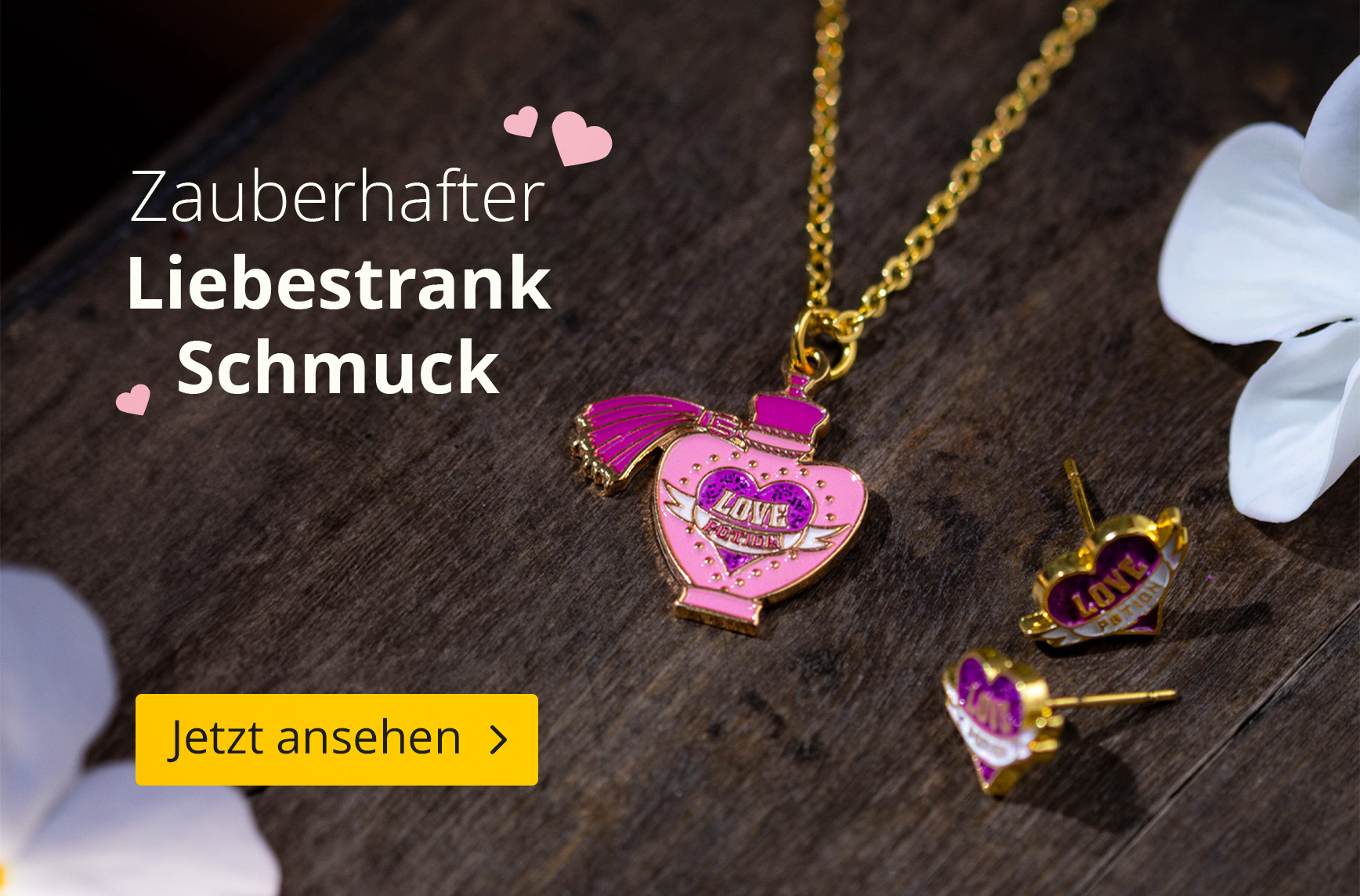 Goldene Kette mit rosa Liebestrank-Anhänger und passenden Herz-Ohrsteckern auf dunklem Holzhintergrund, dekoriert mit weißen Blüten. Text: Zauberhafter Liebestrank Schmuck. Jetzt ansehen.