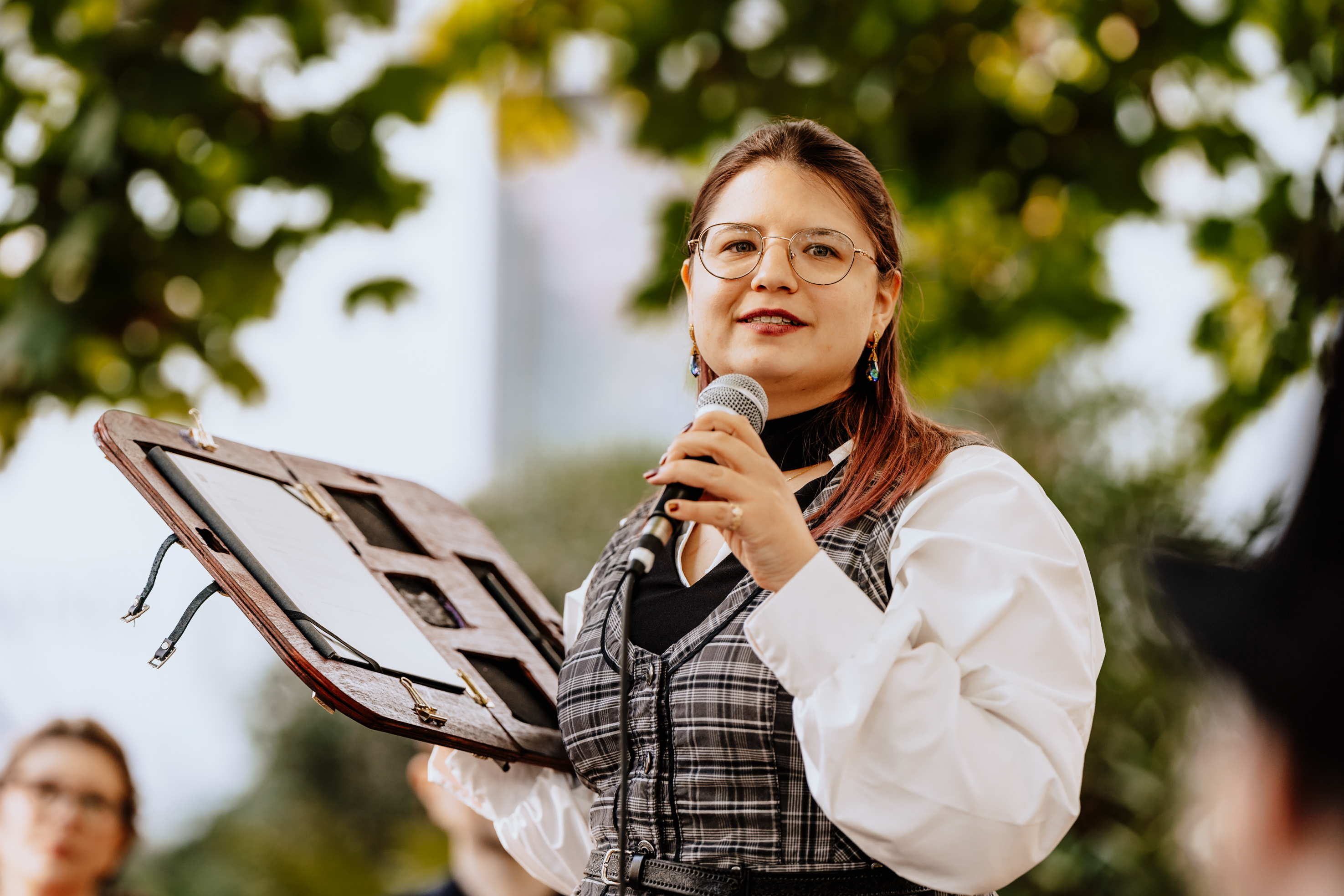 A woman with glasses and a microphone speaks during an outdoor ceremony, holding an open book or folder.