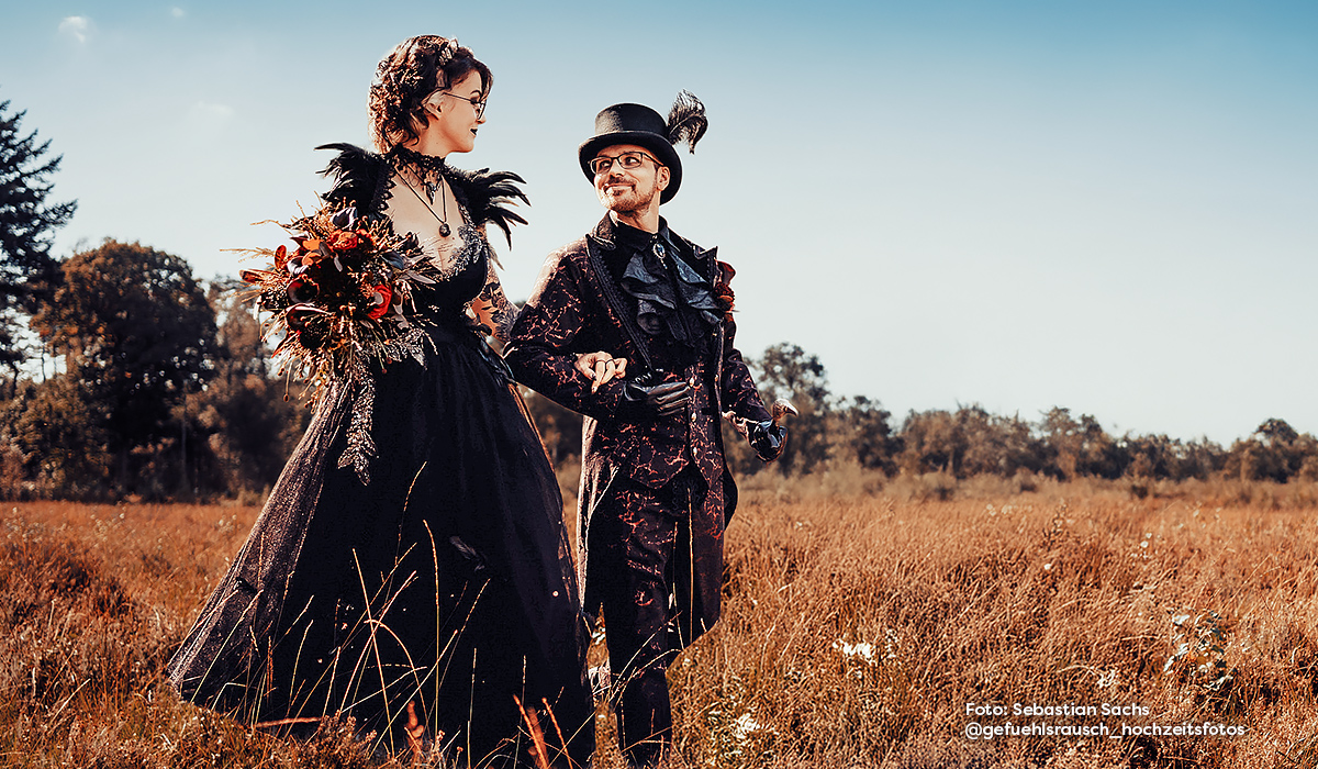 Couple in extravagant, nerdy-inspired wedding outfits posing in a field. She wears a black dress with bouquet, he a suit with top hat. Symbolic image for creative nerdy weddings. Photo: Sebastian Sachs @gefuehlsrausch_hochzeitsfotos