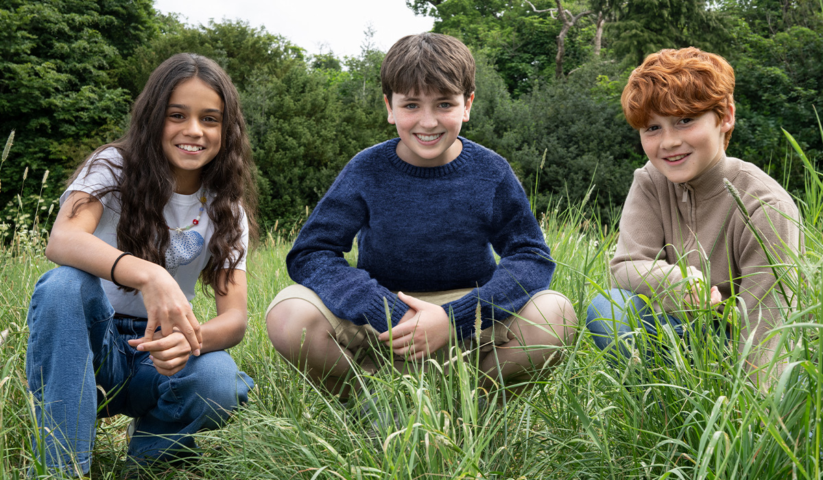 The three main characters from HBO's Harry Potter series, Harry Potter, Hermione Granger and Ron Weasley, are sitting in the tall grass of a green meadow surrounded by trees, looking directly into the camera.
