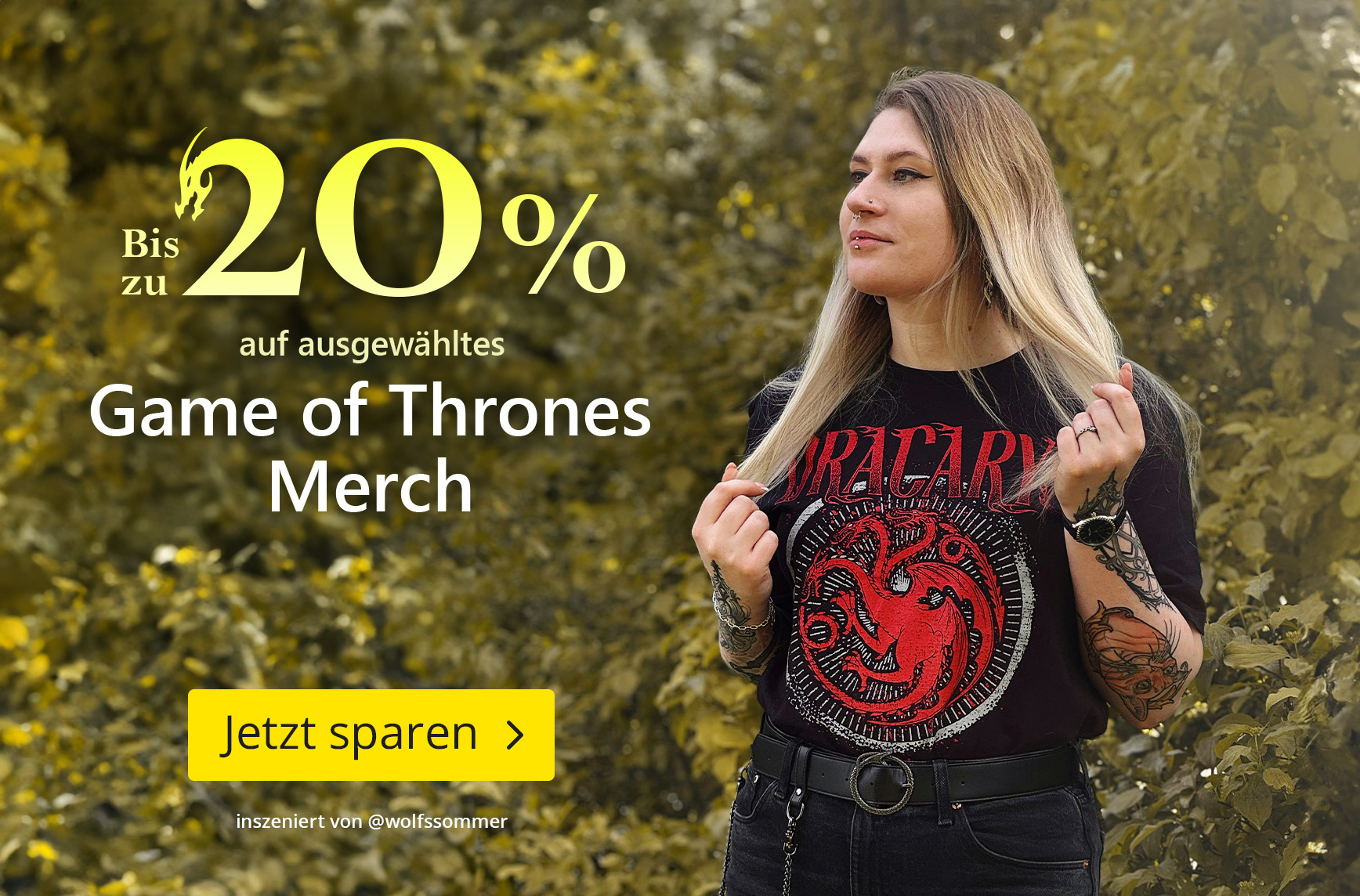 Eine Person trägt ein schwarzes Game of Thrones T-Shirt mit rotem Targaryen-Drachenmotiv und steht vor einem grünen Naturhintergrund. Im Bild wird eine Rabattaktion mit bis zu 20 % auf ausgewähltes Game of Thrones Merch beworben. Text im Bild: „Bis zu 20 