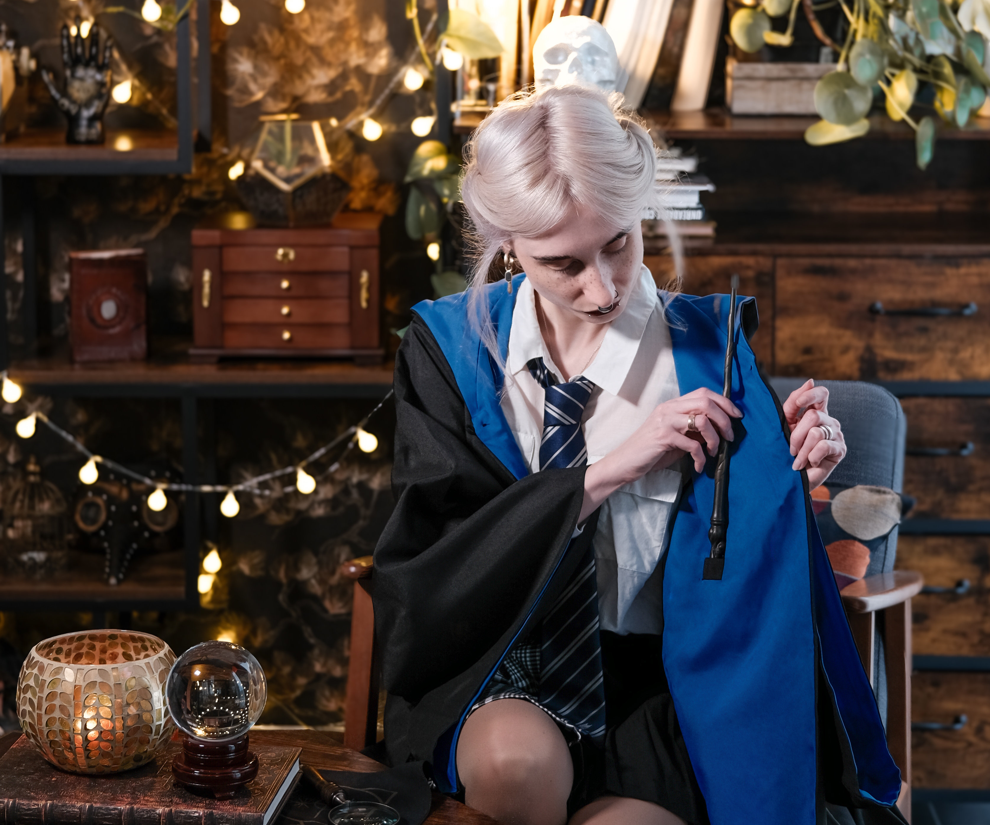 Una persona in cosplay di Harry Potter indossa una tunica Corvonero ed estrae una bacchetta dalla tasca interna. Sullo sfondo: una stanza decorata in stile magico con luci, libri e una sfera di cristallo.