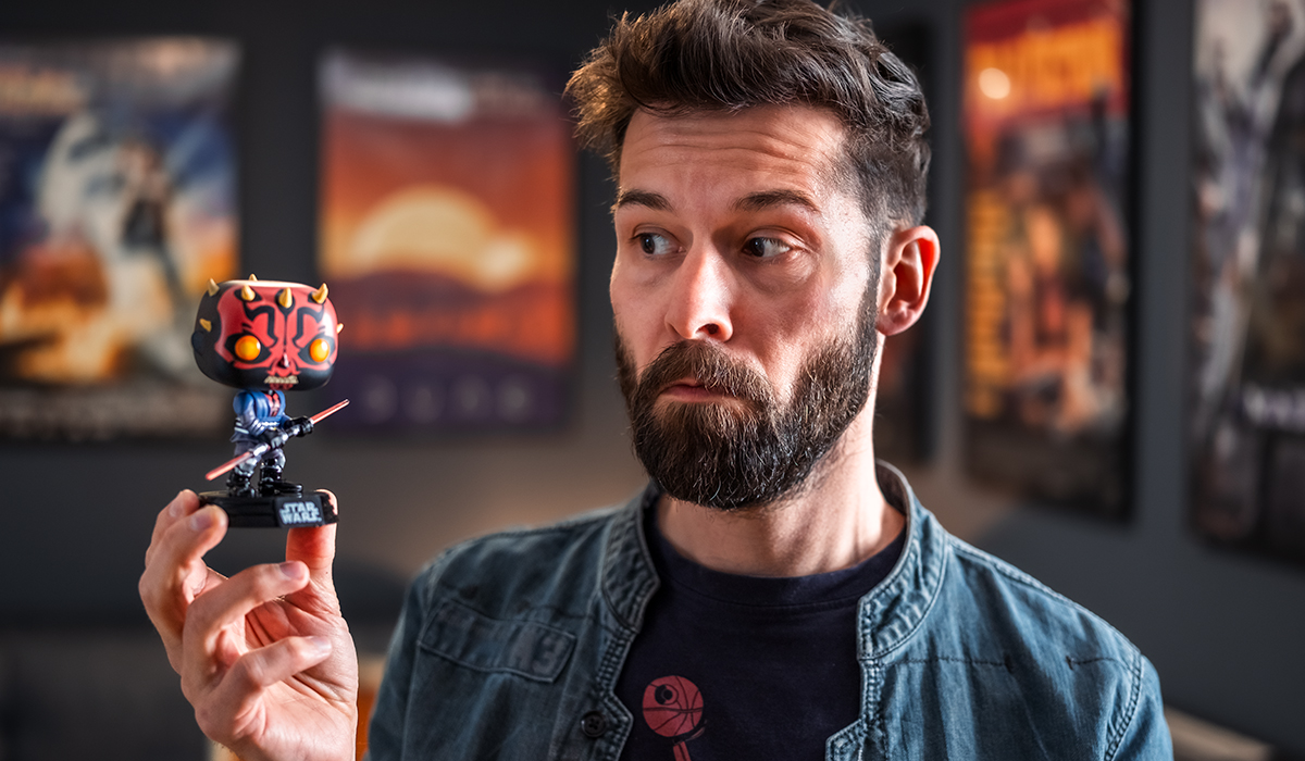 A bearded man looks sceptically at a Darth Maul Funko Pop figure in a mechanical Cyber-Sith design, which he is holding in his hand. In the dimly lit background, framed film posters hang on a dark wall.