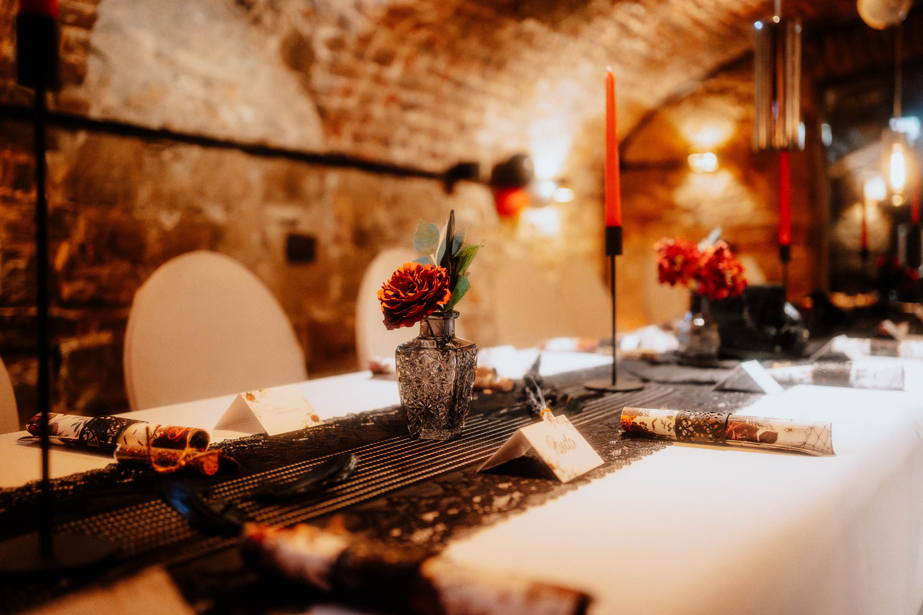 Festively set table in a vaulted cellar, decorated with red candles, black lace runners, and red flowers in glass vases.
