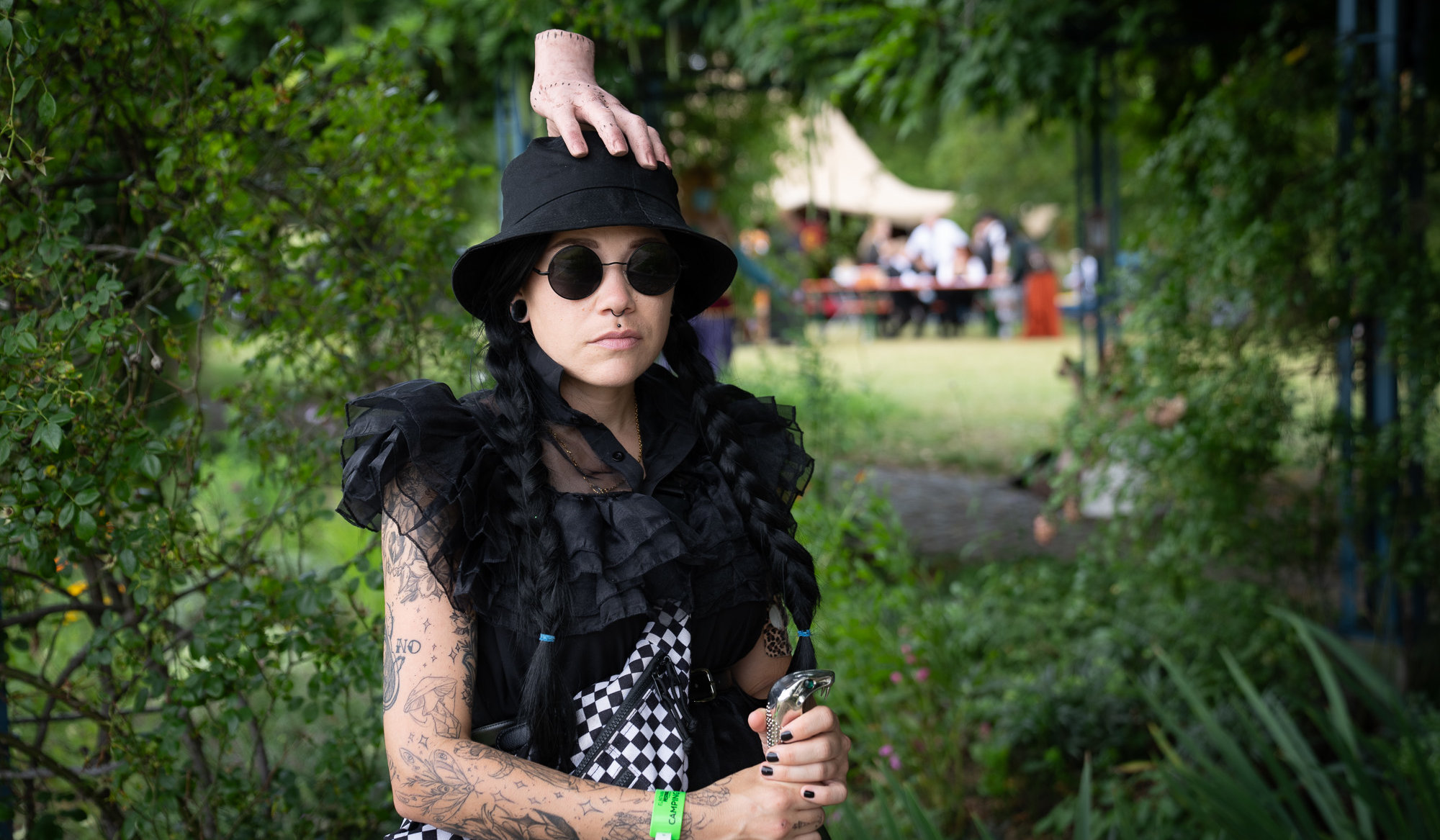 Cosplayerin als Wednesday Addams mit schwarzen Zöpfen, Sonnenbrille und schwarzem Hut, auf dem die „eiskalte Hand“ sitzt. Die Szene spielt draußen auf einem Festivalgelände mit grünem Hintergrund.