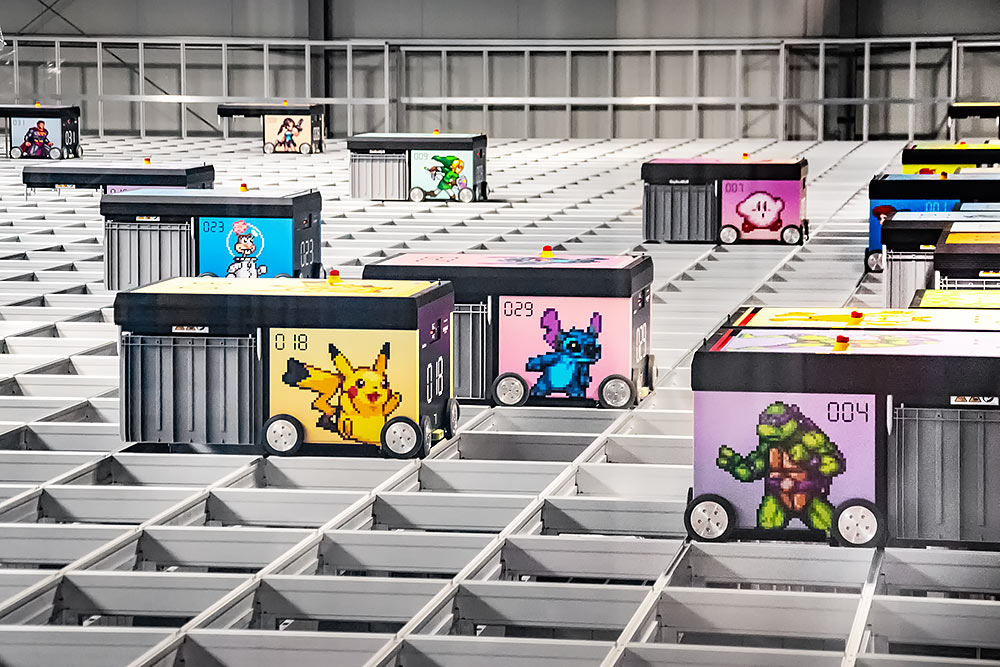 Mehrere AutoStore-Roboter mit nerdigen Pixel-Art-Stickern (u. a. Kirby, Wonder Woman, The Flash und Nyan Cat) bewegen sich über das Lagergitter von Elbenwald.