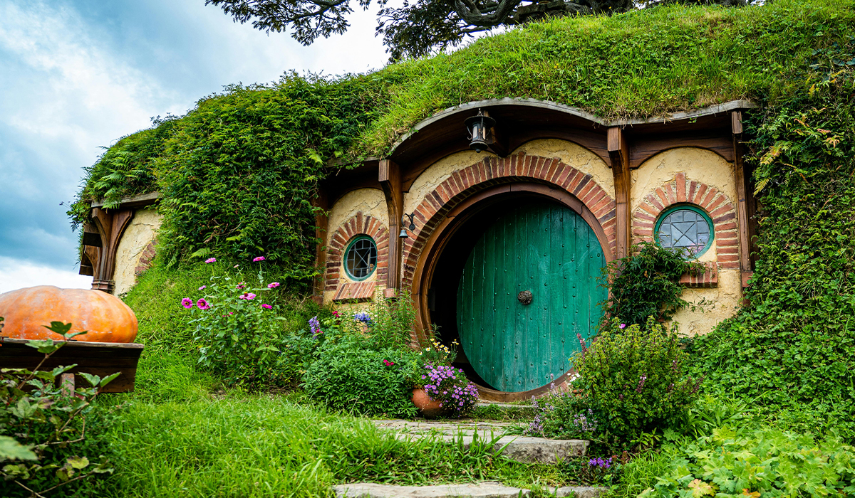 Hobbit hole with a round green door, grass-covered roof and blooming garden in front of the entrance.