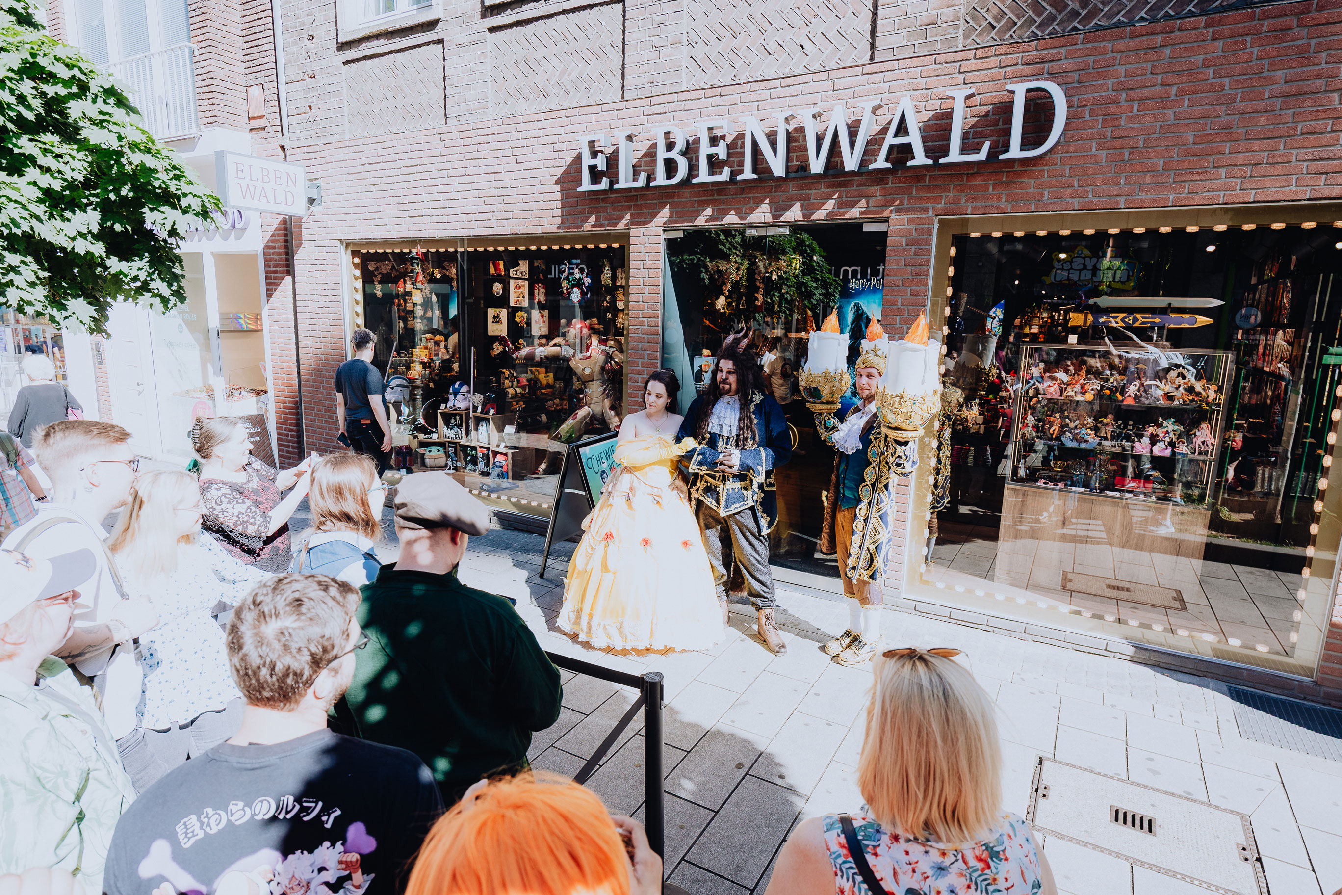 Three cosplayers in Disney costumes stand in front of an Elbenwald store, with a queue of people in front of them.