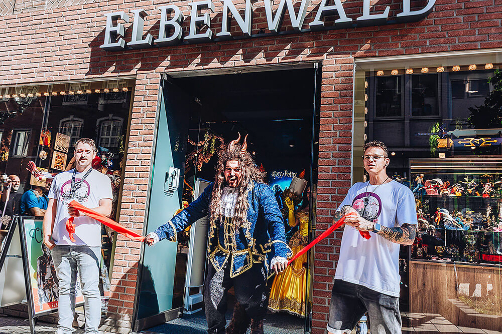 Ein als Biest verkleideter Cosplayer schneidet lachend ein rotes Band vor dem Elbenwald Store durch, flankiert von zwei Mitarbeitenden in weißen T-Shirts. Hinter ihnen sind Schaufenster mit Figuren und Kostümen zu sehen.