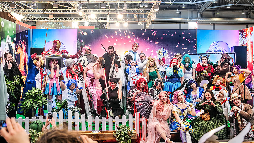 Cosplayer in farbenfrohen Kostümen posieren lachend auf der Bühne des Elbenwald-Stands bei der Leipziger Buchmesse 2025, umgeben von Deko, Pflanzen und einem leuchtenden Hintergrund mit Feuerwerkseffekt.