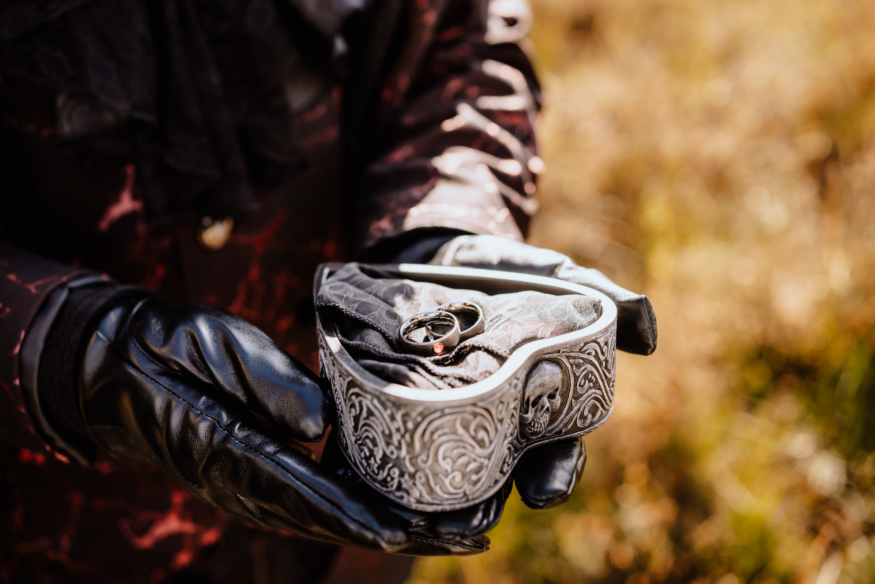 Hands in black leather gloves hold a heart-shaped box with skull engravings, containing two rings on fabric.