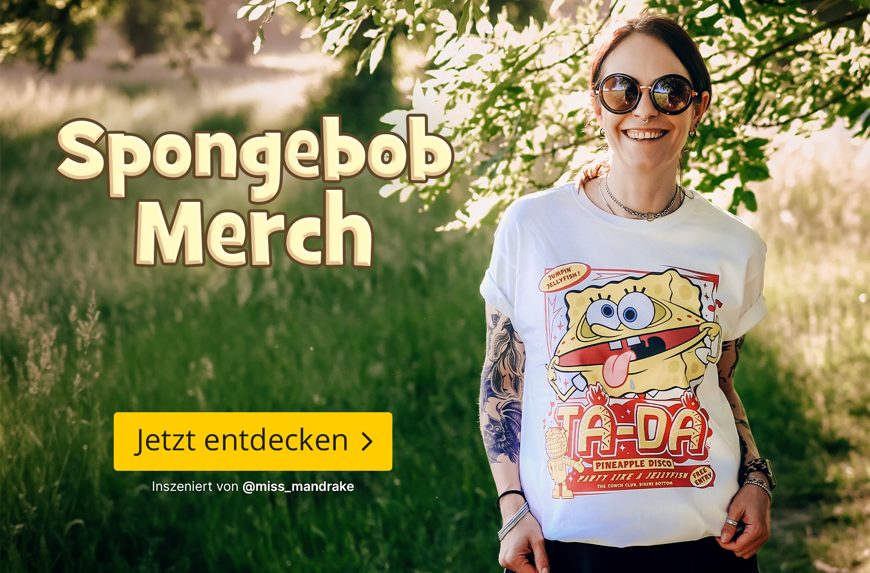 Person mit Sonnenbrille und Tattoos steht auf einem sonnigen Waldweg und trägt ein weißes SpongeBob T-Shirt mit farbenfrohem Motiv. Die Szene wirkt sommerlich und fröhlich. Text im Bild: „Spongebob Merch“ sowie der Button „Jetzt entdecken“.