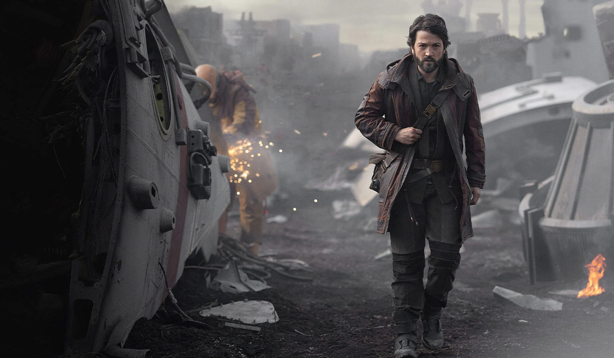 blog-serien-01 Cassian Andor geht entschlossen durch ein trostloses Industriegebiet voller Trümmer und Raumschiffwracks. Im Hintergrund arbeitet ein Mechaniker mit Funkenflug an einem beschädigten Schiff. Die Szene stammt aus der Serie „Star Wars: Andor”.