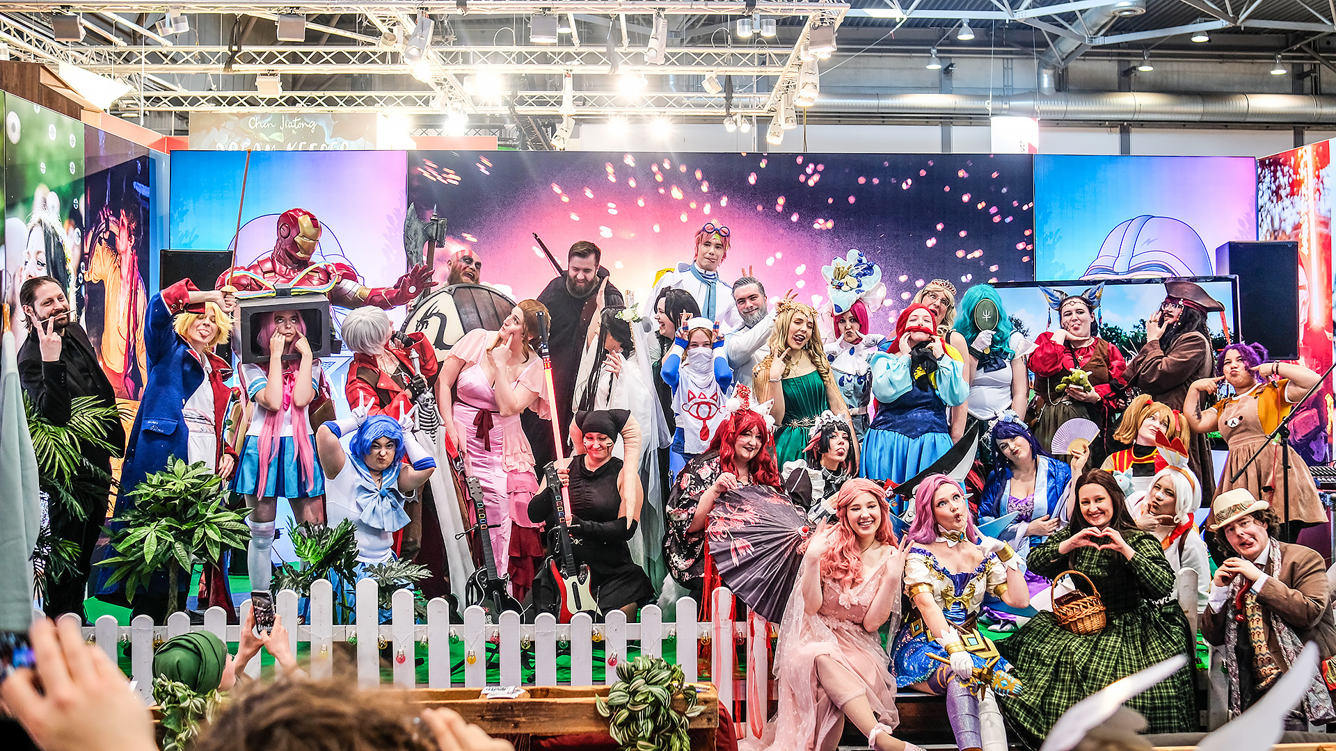 Cosplayers con trajes coloridos posan riendo en el escenario del stand de Elbenwald en la Feria del Libro de Leipzig 2025, rodeados de decoración, plantas y un fondo brillante con efectos de fuegos artificiales.