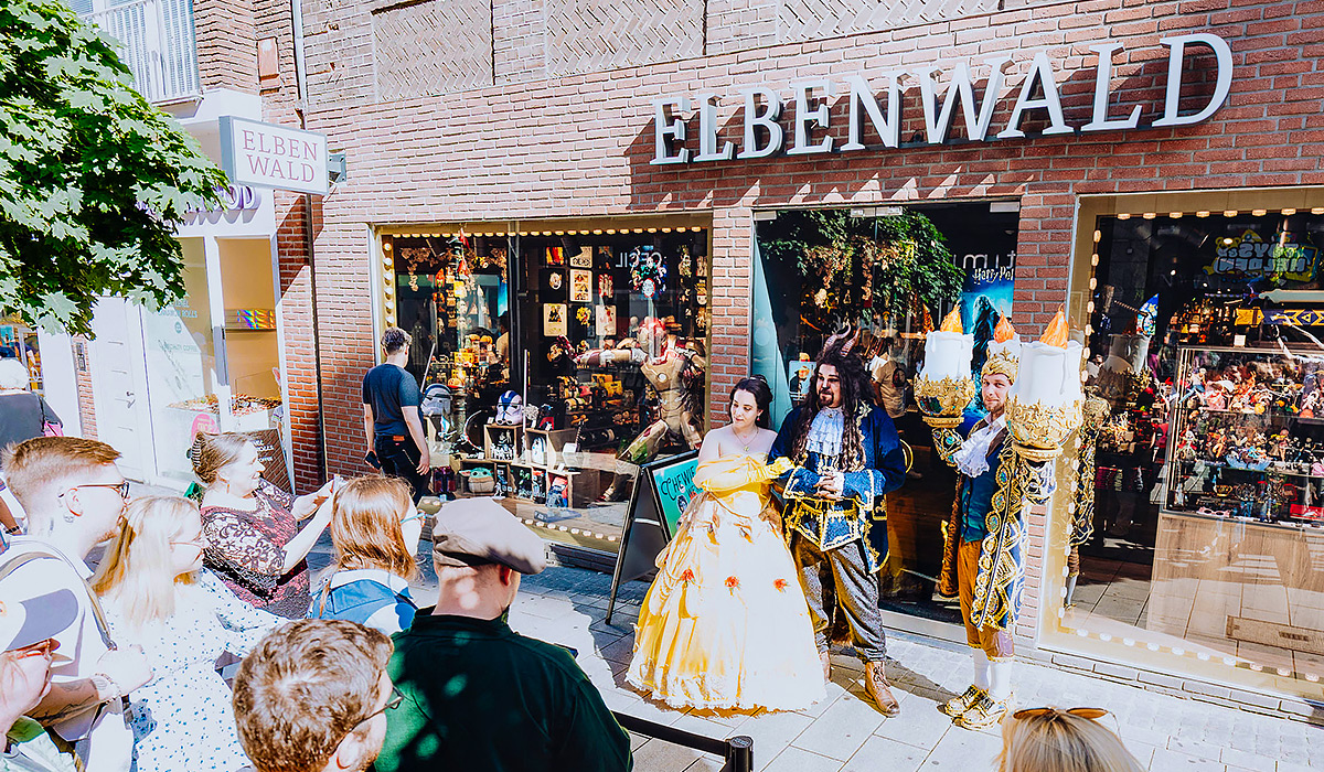 Three cosplayers in Disney costumes stand in front of an Elbenwald store, with a queue of people in front of them. Photo Credit: Rainer Keuenhof