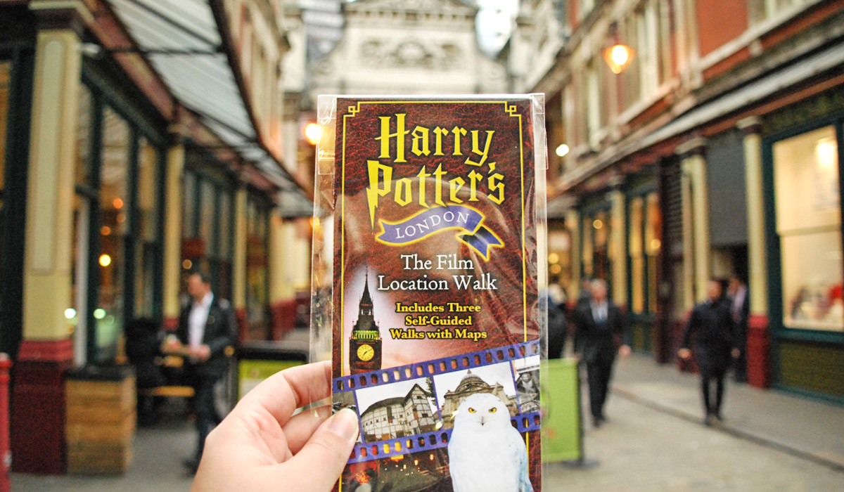 Eine Hand hält einen Flyer zur „Harry Potter’s London – The Film Location Walk“-Tour in die Kamera. Im Hintergrund ist eine überdachte Gasse in London zu sehen – eine bekannte Drehkulisse aus den Filmen.